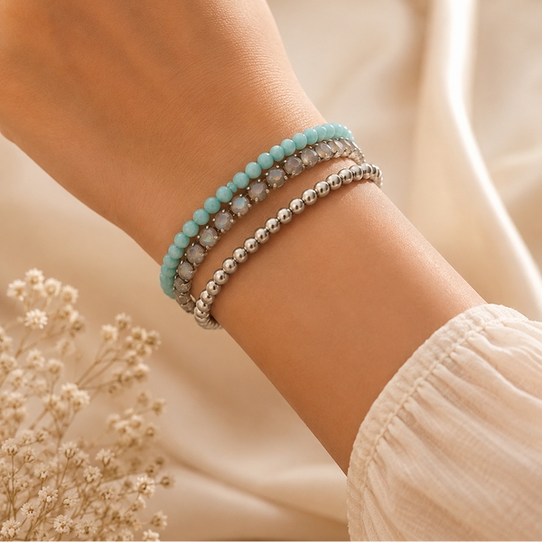 A close-up of a person's wrist wearing three beaded bracelets: a light blue beaded bracelet, a silver beaded bracelet, and a silver bracelet with clear faceted stones. The background is a soft beige fabric with dried white flowers.