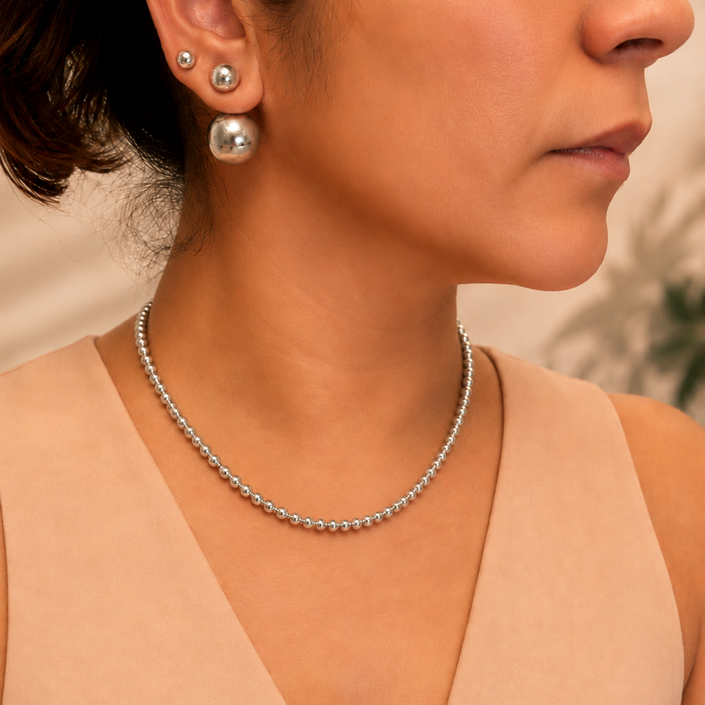 A close-up of a woman wearing a delicate silver beaded chain necklace and matching stud earrings, paired with a beige V-neck top.