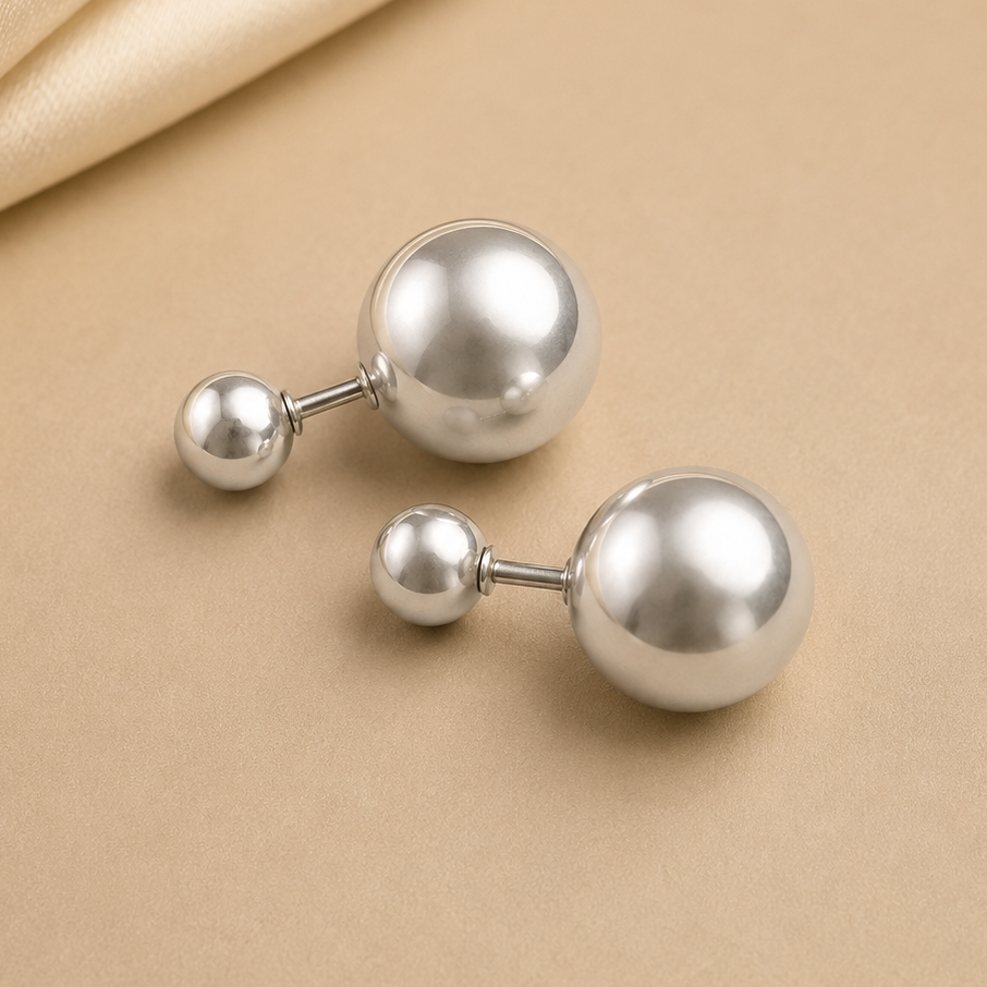 A pair of silver ball stud earrings with smaller matching ball studs, displayed on a beige surface with a draped satin fabric in the top left corner.