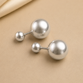 A pair of silver ball stud earrings with smaller matching ball studs, displayed on a beige surface with a draped satin fabric in the top left corner.