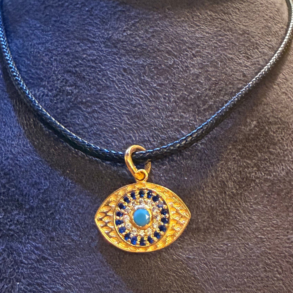 A gold-toned evil eye pendant necklace with a blue center and black surrounding details, displayed on a dark gray fabric jewelry stand. The necklace is on a black cord, set against a warm, amber-colored background with blurred glassware.