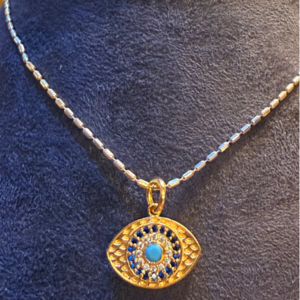 A split comparison of an eye-shaped pendant necklace by Gilbi, showing two styles: a gold pendant with a blue center on a black leather cord, and a gold pendant with a blue center on a sterling silver chain. Both are displayed on blue velvet jewelry busts against a dark gray and light beige background.