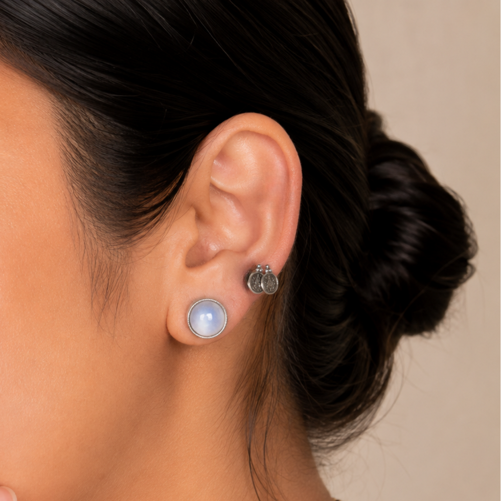 A close-up of a woman wearing a small round light blue pearl stud earring with a silver-toned post, shown against a soft beige background.