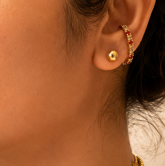 A close-up of a woman wearing a double-layered gold necklace with a red teardrop-shaped pendant, along with a small gold hoop earring.