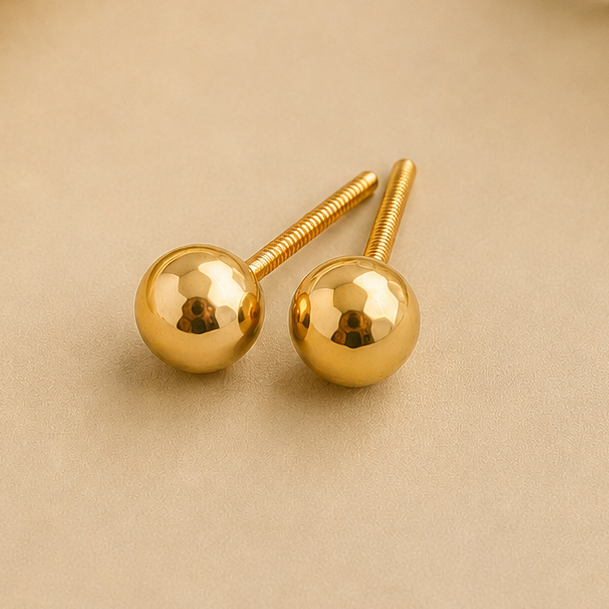 A pair of gold ball stud earrings with straight posts, displayed on a beige surface with a draped cream satin fabric and small white flowers in the background.