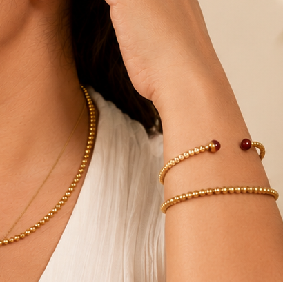 A woman wearing a white sleeveless top and gold jewelry, including a necklace with a small dark red pendant and matching bracelets, in a warm, sunlit indoor setting.