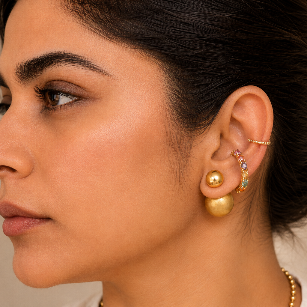 A close-up side profile of a woman wearing gold ear piercings, including a large gold hoop earring, a gold stud, a gold helix band, and a small multicolor stone-set cartilage hoop, against a warm beige background with soft leaf shadows.