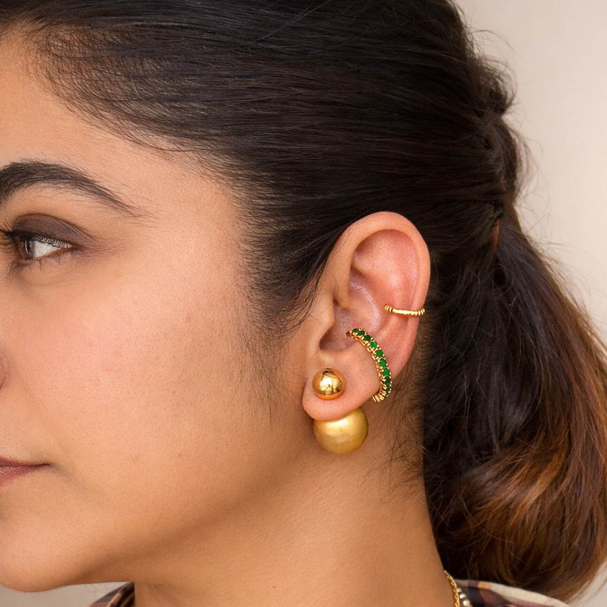Close-up of a woman's ear wearing multiple gold-toned earrings, including a large gold disc earring, a curved gold hoop with green stones, and a thin gold ear cuff, against a soft beige background.