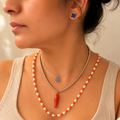 A close-up of a woman wearing layered necklaces, including a coral and white pearl beaded necklace with a red tassel pendant, a silver beaded necklace with a blue evil eye pendant, and a silver chain necklace, along with a matching coral and white pearl hoop earring.
