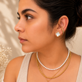 A woman in profile wearing layered necklaces and earrings, featuring a white pearl strand, a gold beaded chain, a gold chain with a red teardrop pendant, and a gold hoop with a white flower stud earring.