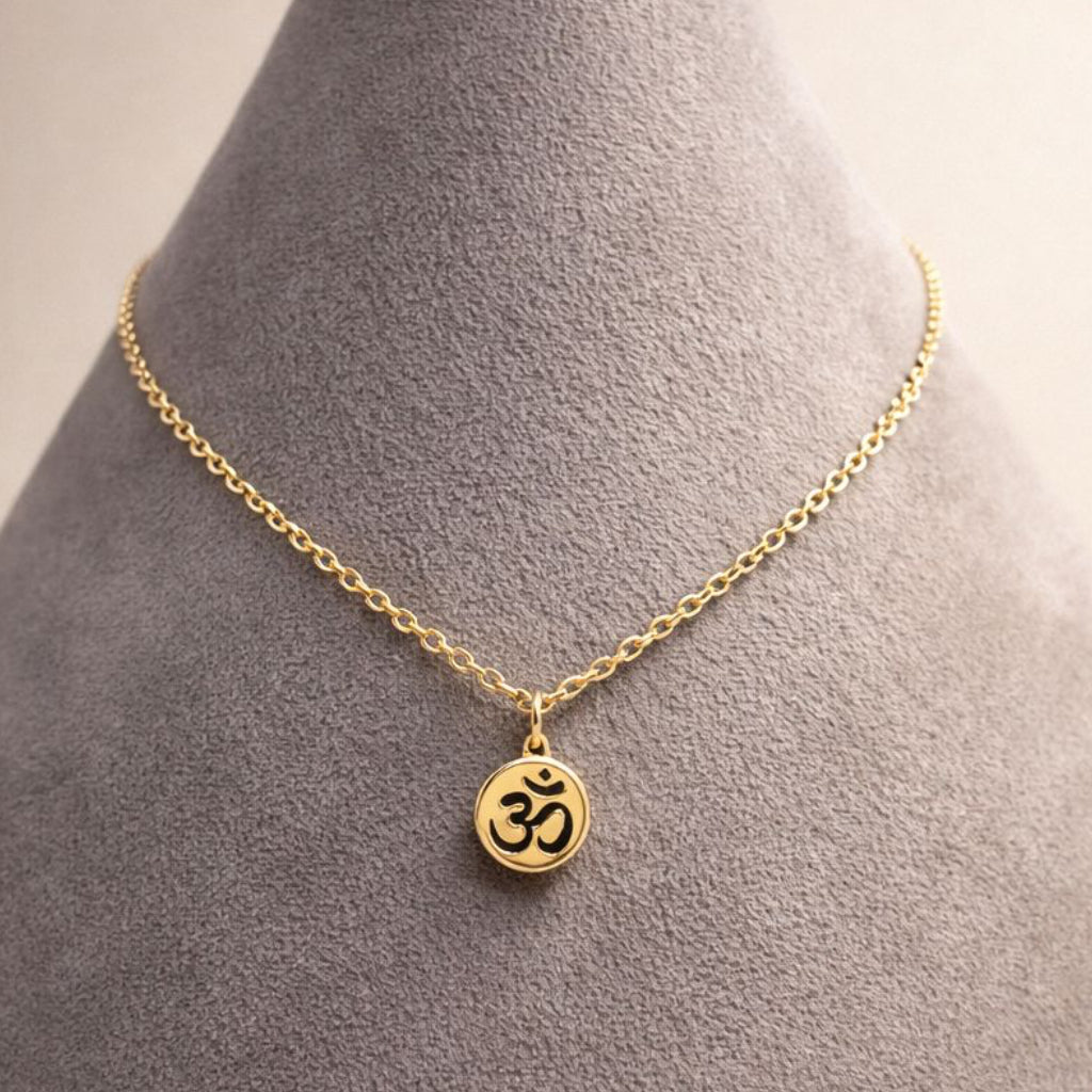 A delicate gold chain necklace with a small circular pendant featuring a black Om symbol, displayed on a gray velvet jewelry bust against a light beige background.