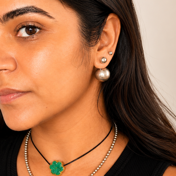 A woman wearing layered necklaces, including a black cord necklace with a green clover pendant and a silver beaded chain, along with pearl stud earrings, against a plain beige background.