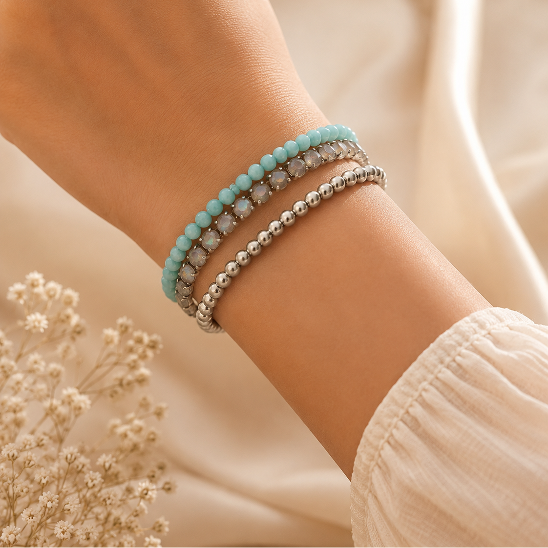 A close-up of a person's wrist wearing three beaded bracelets: a light blue beaded bracelet, a silver beaded bracelet, and a silver bracelet with clear faceted stones. The background is a soft beige fabric with dried white flowers.