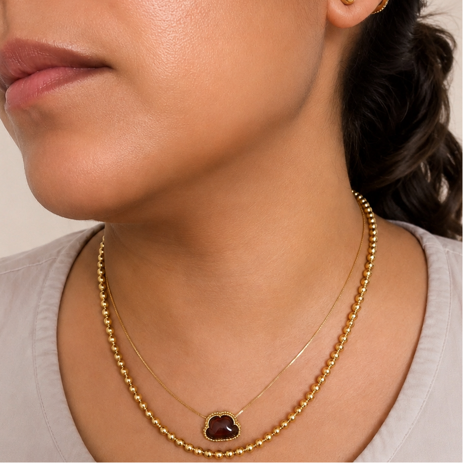 Close-up of a woman wearing a layered necklace set featuring a gold beaded chain and a delicate chain with a small heart-shaped dark red gemstone pendant.