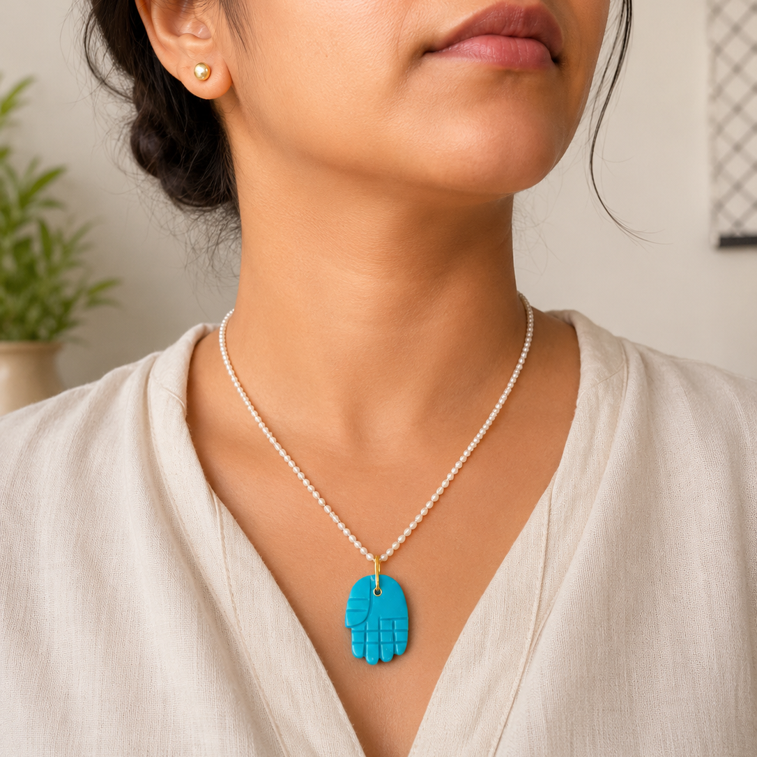 A close-up of a woman wearing a turquoise hand-shaped pendant necklace on a delicate white beaded chain, paired with a small gold stud earring.