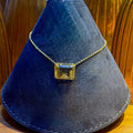 A gold chain necklace with a square-cut yellow gemstone pendant, displayed on a dark blue velvet jewelry cone stand.